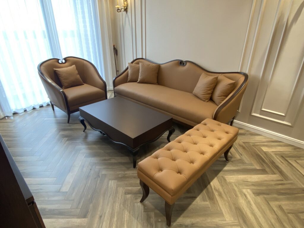 Bộ sofa Daphne bọc tông nâu bò, bàn trà gỗ tối và đôn chần nút trong phòng khách sàn gỗ xương cá