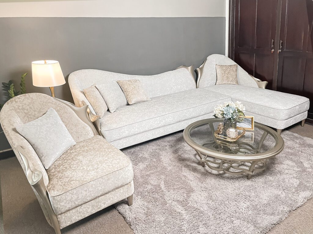 Armchair Di Angelo tân cổ điển bọc vải hoa văn kem, tựa lưng cuộn lệch một bên, đặt cạnh sofa góc và bàn trà kính tròn
